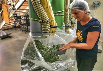 Eine Mitarbeiterin entnimmt mit einer Futterschaufel Kräuter aus einem großen Sack, in den eine Röhre hineinführt.  | © SONNENTOR